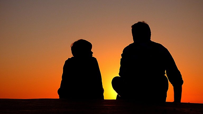 Kuvassa miehen ja lapsen hahmot istuvat auringonlaskua katsellen
