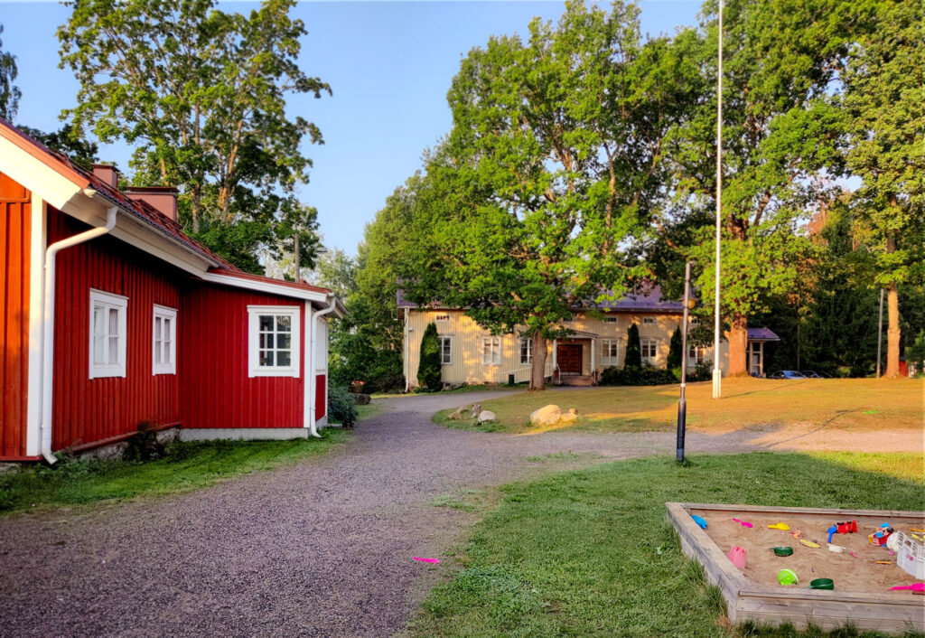 pihapiiri, jossa näkyy punainen talo. taempana keltainan talo puun takana. edessä hiekkalaatikko