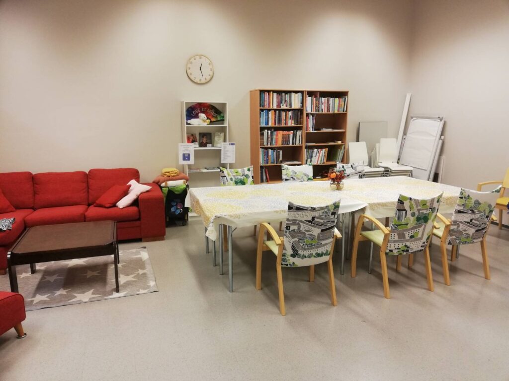 Yleiskuva Simpukan toimiston kahvittelutilasta. Vasemmalla nojatuoli jalkajakkaroineen, matala pöytä ja kulmasohva. Oikealla iso kahvipöytä, jonka ympärillä on 8 tuolia. Lisää tuoleja pöydän oikeanpuoleisessa päädyssä. Pöydän takana seinää vasten on pari kirjahyllyä.