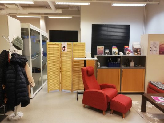 Yleiskuva Simpukan toimistolta henkilökunnan työtilan suuntaan. Vasemmalla naulakot, oikealla nojatuoli ja matala sohvapöytä. Suoraan edessä kaappeja ja henkilökunnan puolelle kulun estävä kevyt sälesermi.