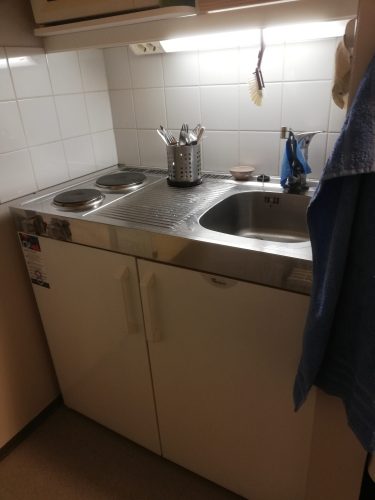 Pieni keittiö. Vasemmalla käytöstä poistettu kaksilevyinen keittolevy, keskellä kapea pöytätaso ja oikealla lavuaari. Keittiön tiskirätti roikkuu seinällä lavuaarin oikealla puolella, ja sen vieressä käsipyyhe. Keittiön astiapyyhe roikkuu seinällä lavuaarin ja keittolevyn vasemmalla puolella.