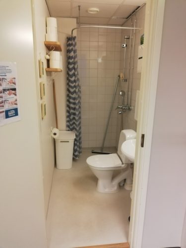 Simpukan toimiston vessa ovelta sisään katsottuna. Vessa on kapea, kolmisen metriä pitkä tila. Käsipesuallas on välittömästi oven oikealla puolella, ja käsipyyhe roikkuu altaan oikealla puolella seinällä. Pytty on altaan jälkeen oikealla, ja vessapaperirulla pytyllä istuessa suoraan edessä vastakkaisella seinällä. Pytyn jälkeen tilan perällä on pyykkikori ja suihku.