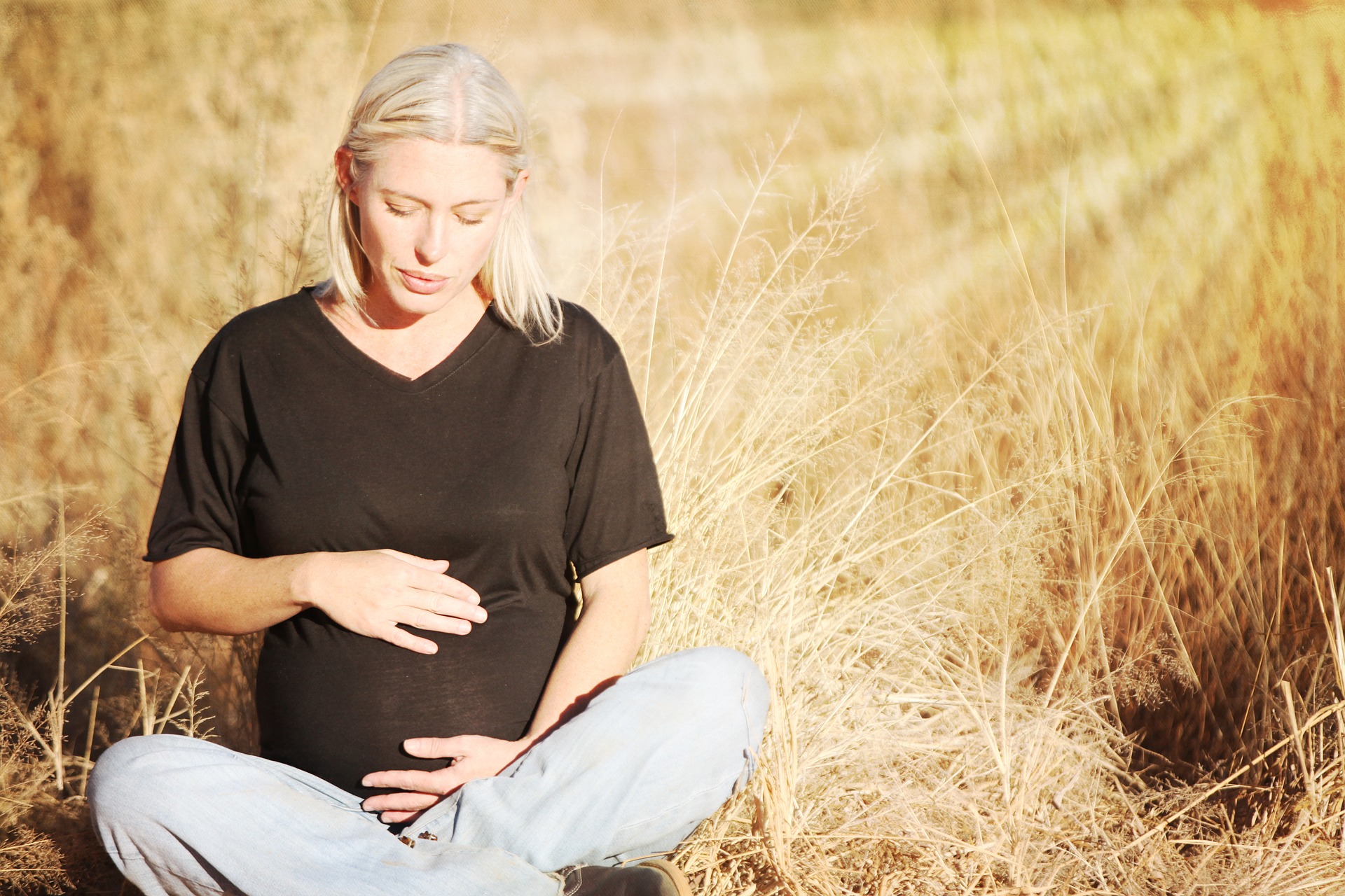 Raskaana oleva nainen istuu heinikossa ja pitele käsiään raskausmahan päällä.