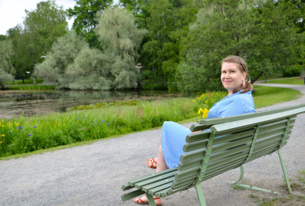 Nainen istuu valkoisella puiston penkillä, katsoo kameraan ja hymyilee, taustalla näkyy lampi ja vihreää luontoa.