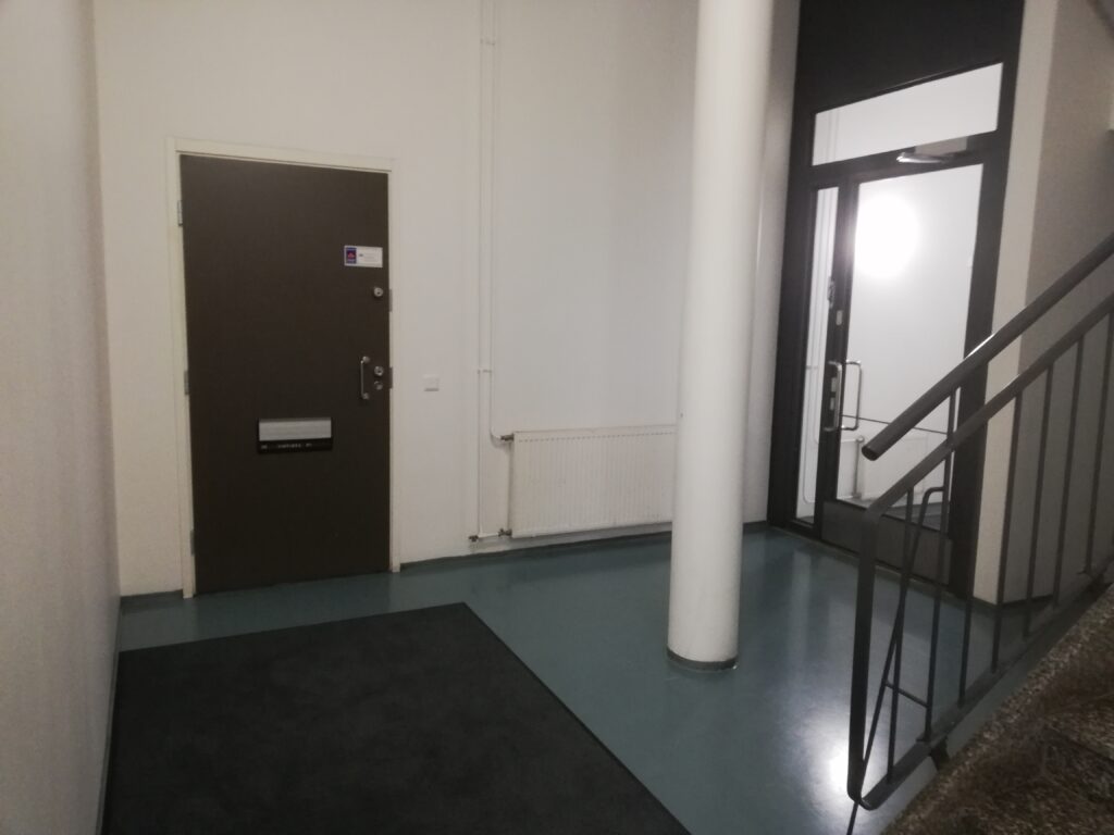 Simpukan toimiston ovi kerrostalon ala-aulassa vasemmalla. Aulaan tullessa suoraan edessä hissi ja rappukäytävä yläkertaan. Simpukan oven ja rappukäytävän välissä viistosti etuvasemmalla kivipylväs ja sen takana ovi Massun parkki -parkkihalliin.